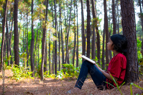 Little girl sitting under pine tree over pine forest. In she hand have notebook , she look  learning nature for write something to notebook. Concepts of learn beyond classroom.