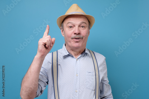 Senior hispanic man in summer hat looking up and pointing his finger. I have an Idea concept. Studio shot on blue wall. © Viktor Koldunov