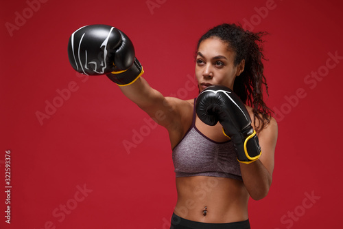 Confident young african american sports fitness boxer woman in sportswear working out isolated on red wall background. Sport exercise healthy lifestyle concept. Make boxing exercises in boxing gloves © ViDi Studio