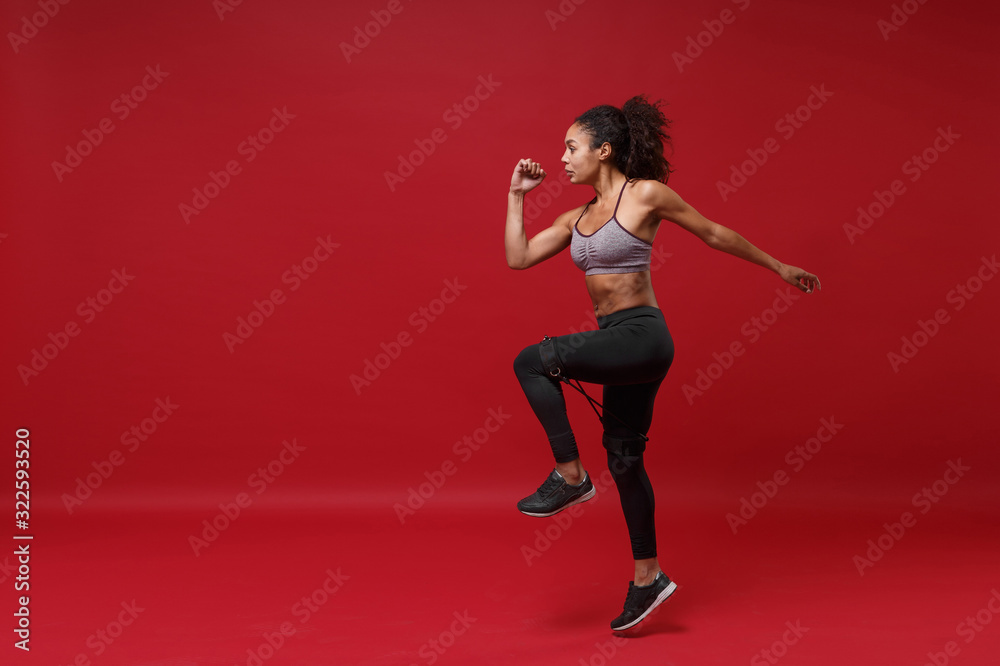 Side view of young african american sports fitness woman in sportswear working out isolated on red background in studio. Sport exercises healthy lifestyle concept. Doing exercises with elastic band.