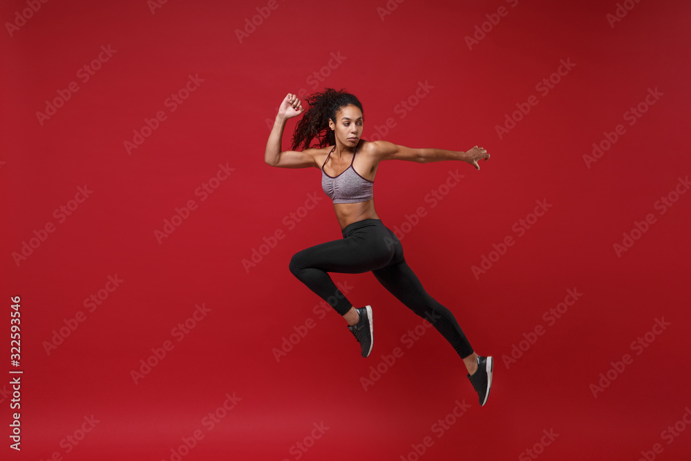 Side view of young african american fitness woman in sportswear posing working out isolated on red wall background studio portrait. Sport exercises healthy lifestyle concept. Jumping like running.