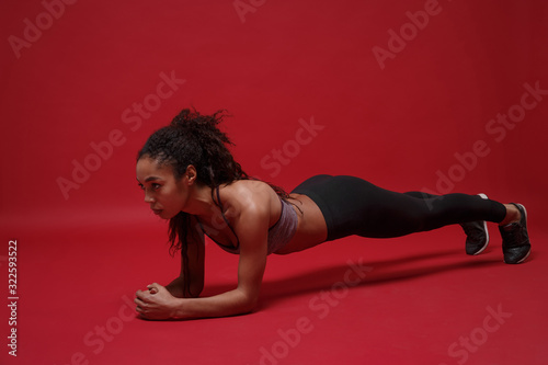 Strong beautiful young african american sports fitness woman in sportswear working out isolated on red background studio portrait. Sport exercises healthy lifestyle concept. Doing plank exercises