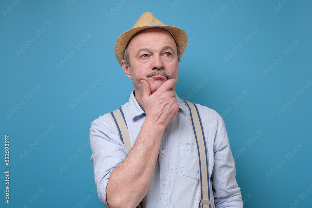 Senior man in summer hat thinking and rubbing a chin trying to make a right solution. Studio shot on blue wall