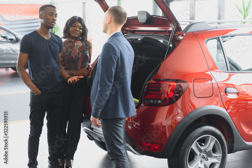African american family at car dealership. Salesman is showing trunk of new red car. © Serhii