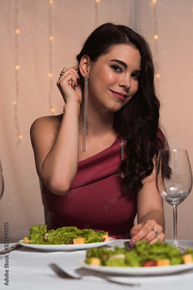 smiling, elegant girl touching hair while sitting at served table in restaurant