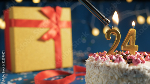 Photography White birthday cake number 24 golden candles burning by lighter, blue background with lights and gift yellow box tied up with red ribbon