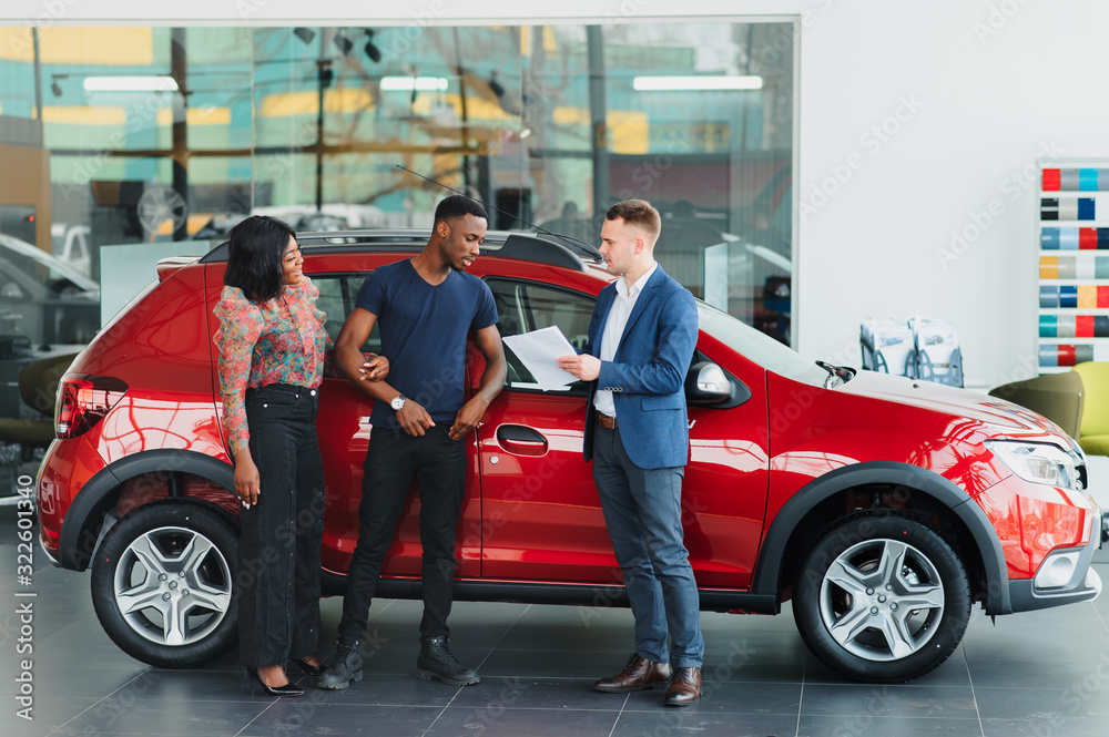 Naklejka premium happy african couple choosing luxury car at vehicle dealership looking at the interior