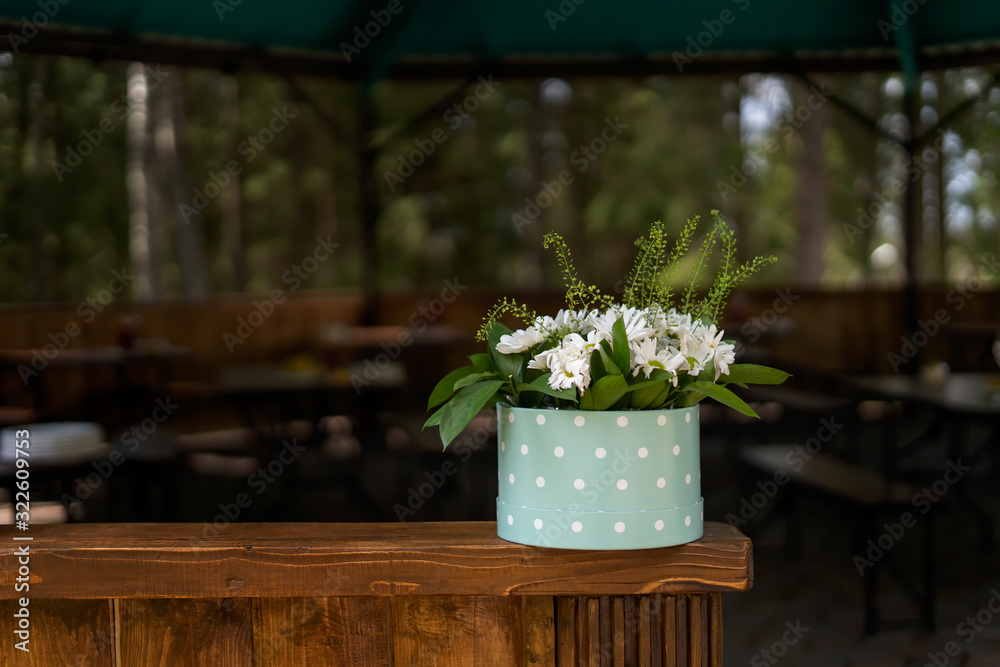 Large bouquet of daisies in a round cardboard box, stands on a wooden railing.