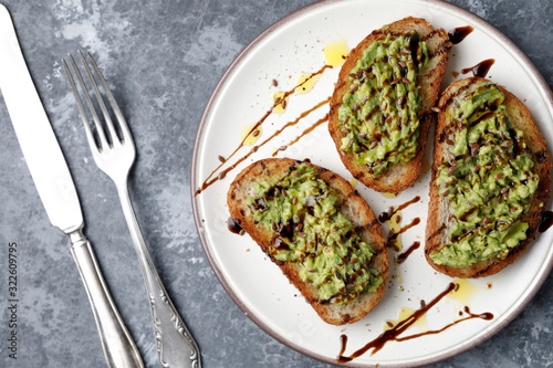 A freshly made minced avocado toast with balsamico dressing, linseed or flaxseed and oil on a white plate, on a gray background and silver fork and knife