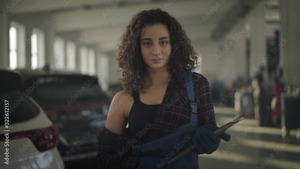 Middle shot of young female auto mechanic posing in car repair shop. Beautiful brunette Caucasian woman with curly hair and brown eyes standing in sunrays and looking at camera. Lifestyle, profession. Middle shot of young female auto mechanic posing in car repair shop. Beautiful brunette Caucasian woman with curly hair and brown eyes standing in sunrays and looking at camera. Lifestyle, profession.