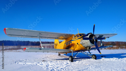 Fototapeta Naklejka Na Ścianę i Meble -  A yellow old biplane plane is parked on a winter airfield against a background of bright blue sky and white snow with the engine running and smoke