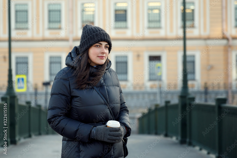 The girl in the jacket with coffee. The girl in a warm jacket on the street smiling and holding a Cup of coffee The girl in the jacket with coffee. The girl in a warm jacket on the street smiling and holding a Cup of coffee