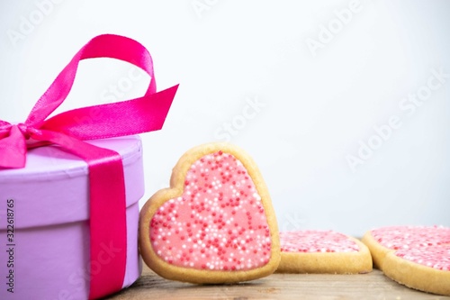Close up of a heart-shaped cookie with pink, white and red sprinkles laying on a present box with a fuchsia ribbon on a wooden base and a white background. Love concept