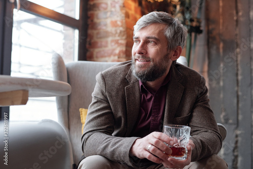 Wall Mural Positive dreamy middle-aged bearded man in brown jacket sitting in armchair and