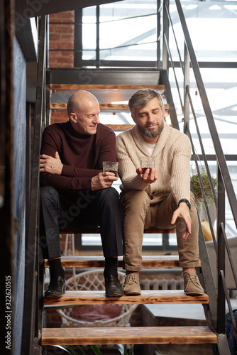 Handsome bearded man with whisky glass sitting on stairs and sharing life story with friend