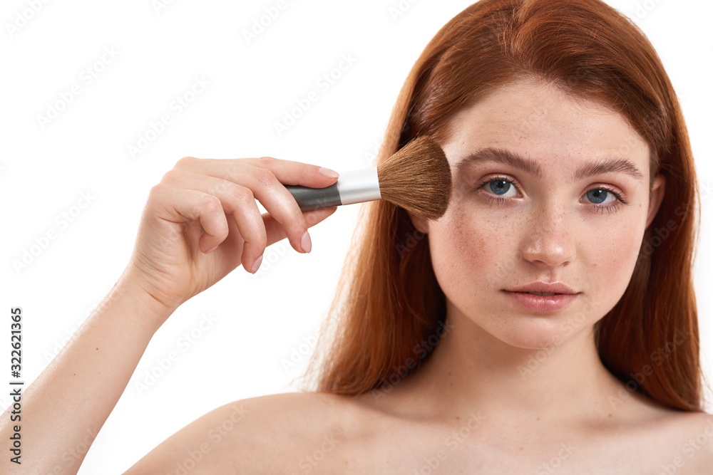 Putting some makeup. Portrait of half naked cute red-haired woman with freckles holding makeup brush and looking at camera while standing in studio against white background Putting some makeup. Portrait of half naked cute red-haired woman with freckles holding makeup brush and looking at camera while standing in studio against white background
