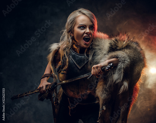 Fotografie, Obraz Fantasy woman knight wearing cuirass and fur, holding a sword and rushes into battle with a furious cry