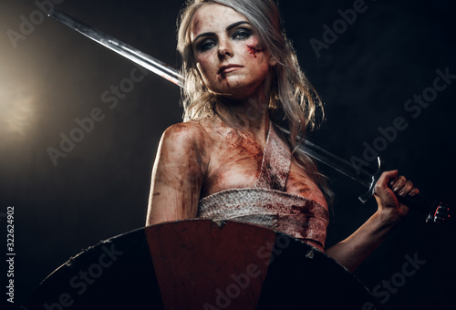 Fantasy woman warrior wearing rag cloth stained with blood and mud, holding sword and shield. Studio photo on a dark background. Cosplayer as Ciri from The Witcher