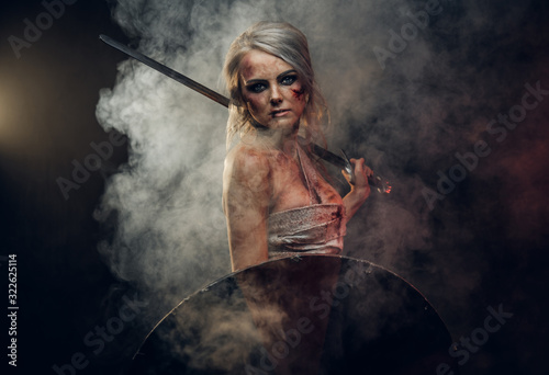 Woman warrior wearing rag cloth stained with blood and mud posing with a sword and shield. Studio photo on a dark background with smoke. Cosplayer as Ciri from The Witcher