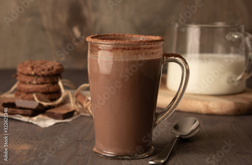 Transparent cup of hot chocolate with cinnamon and milk on a wooden table with oatmeal cookies and chocolate.