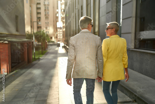 Back view of stylish middle-aged couple holding hands and looking at each other while walking together through the city street © Svitlana