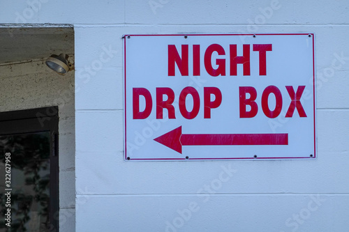 White cinder block wall with a white sign with red letters that say night drop box with an arrow