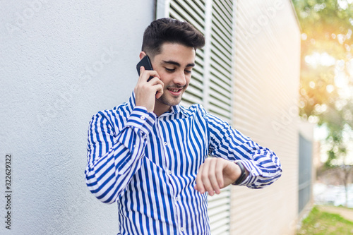 Young man standing outdoors while looking his wrist watch and talking by phone