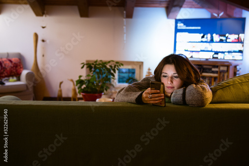 Cute young woman looking at phone in her hand shopping online while sitting on the couch in beautiful interior living room, television on background, relaxation and buying online icon 