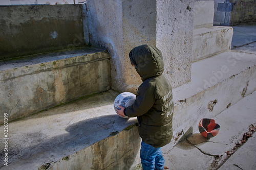 Boy in blue trousers and green jacket playing ball on cement stairs.