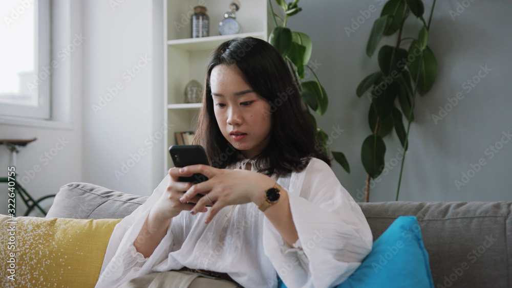 Serious young Chinese brunette using mobile phone, browsing on Internet. Cute girl sitting on sofa, chatting. Indoors. Modern apartment. Technology. Indoors Serious young Chinese brunette using mobile phone, browsing on Internet. Cute girl sitting on sofa, chatting. Indoors. Modern apartment. Technology. Indoors