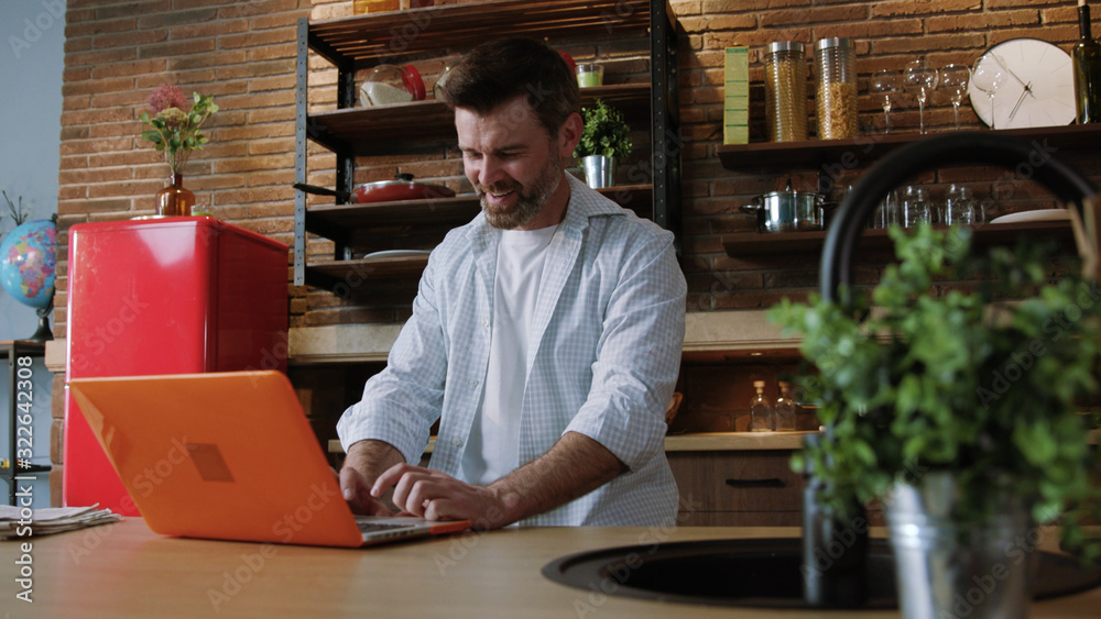 Fototapeta premium Portrait of a sociable man opening his laptop checking social networks and writing his friends online standing in the cozy kitchen at home.