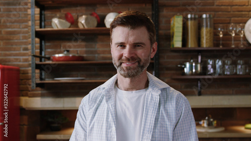 Portrait of a mature handsome bearded man in white shirt standing by kitchen table and smiling of joy. Happy people at home concept. © Fractal Pictures