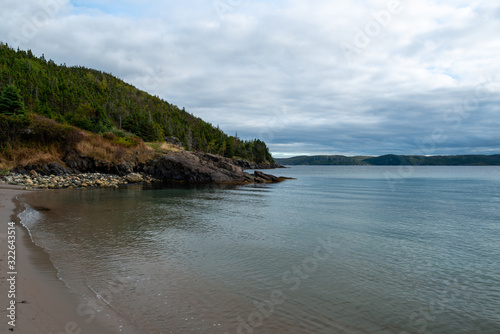 A sandy beach in a small narrow bay. There's nobody on the beach, the water is calm with blue reflections of the sky. A mountain covered in trees is in the background under a cloudy blue evening sky. 