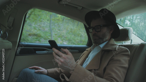 Profile of concentrated mature man in glasses checking business news on a smartphone inside a car. Rich confident businessman texting with a partner while driving in a luxury taxi. © Fractal Pictures
