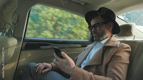 Portrait of Englishman wearing hat and glasses reading news on a mobile phone looking at screen sitting in the back of the luxury car driving in the city. © Fractal Pictures