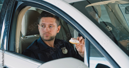 Friendly police officers talking sitting inside the patrol car eating doughnuts together. Two policemen on duty having a tasty break. © Fractal Pictures