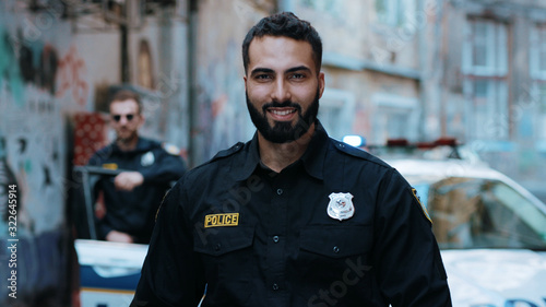 Strong smiling young man cops stand near patrol car look at camera enforcement officer police uniform auto safety security communication control policeman close up slow motion
