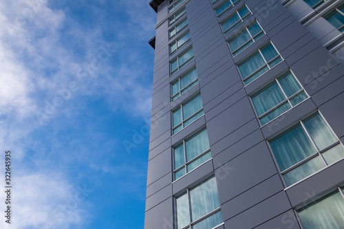The corner exterior wall of a tall building with windows that are reflecting the blue and cloudy sky. The wall is made of grey metal panels.  The blue sky with thin white clouds are in the background.