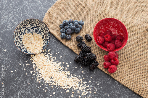 Rolled oat with a mix of Red raspberries falling from a ceramic plate and a bunch of blackberries and blueberries in a rattan and black background. Stylish superfood photography. Healthy breakfast