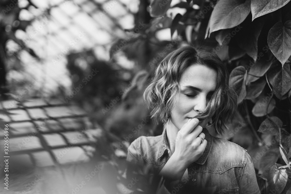 Young beautiful caucasian woman in glass greenhouse among colorful greenery leaves and flowers. Art portrait of a girl wearing a shirt. Gardener, nature lover, inspiration concept. Young beautiful caucasian woman in glass greenhouse among colorful greenery leaves and flowers. Art portrait of a girl wearing a shirt. Gardener, nature lover, inspiration concept.