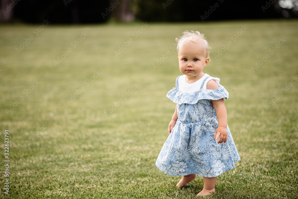 Adorable blue-eyed baby girl in a beautiful blue dress stands barefoot on the grass Adorable blue-eyed baby girl in a beautiful blue dress stands barefoot on the grass