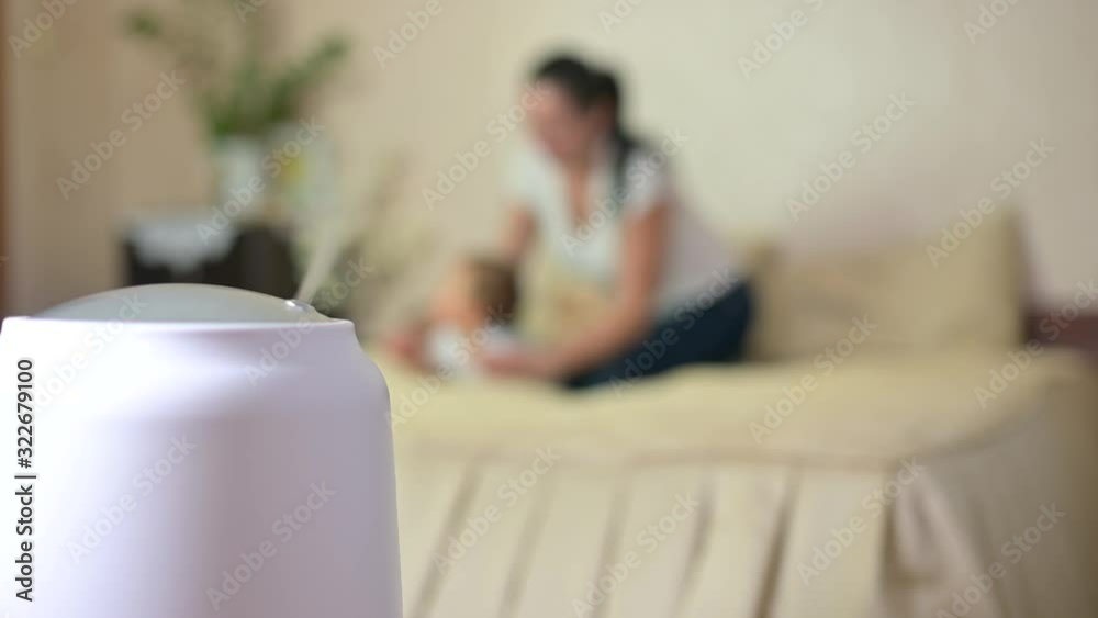 Steam from a working humidifier with ionization of the air in the room, on a blurred background, mom plays with the baby on the bed. humidifier works. Climatic device. Steam from a working humidifier with ionization of the air in the room, on a blurred background, mom plays with the baby on the bed. humidifier works. Climatic device.