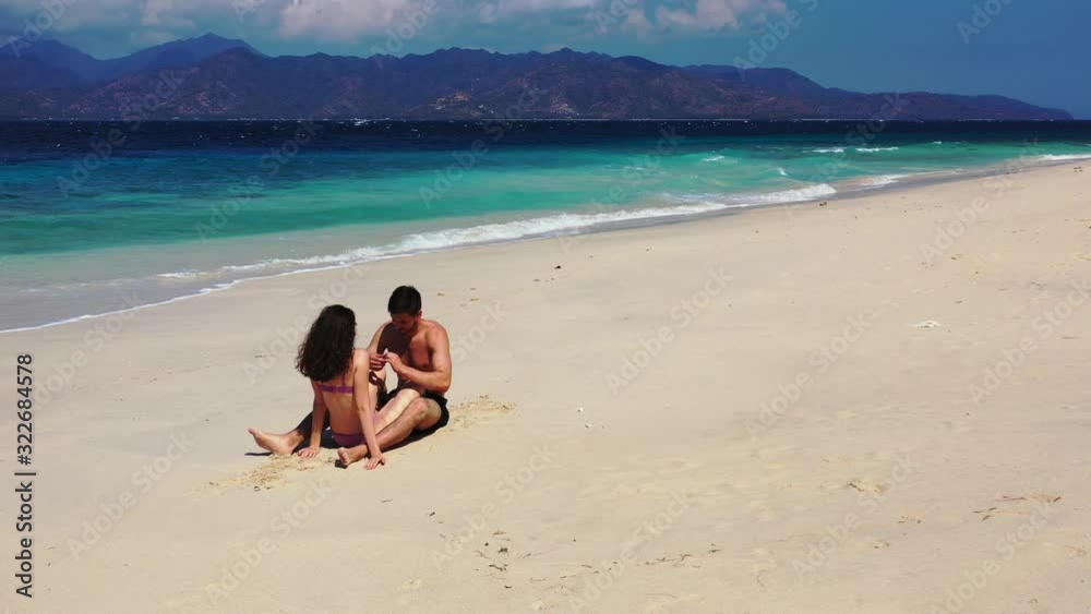 Happy young couple sitting and relaxing while sunbaking on a beautiful beach with mountains in the distance Happy young couple sitting and relaxing while sunbaking on a beautiful beach with mountains in the distance