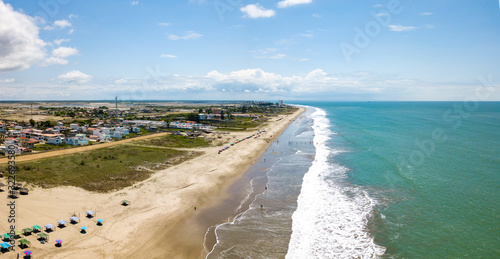 Aerial drone view of the beach, tourists, sea and waves splashing in the coast of the town Playas General Villamil, Ecuador. Sunny day. Beach houses and the horizon with some clouds in background.