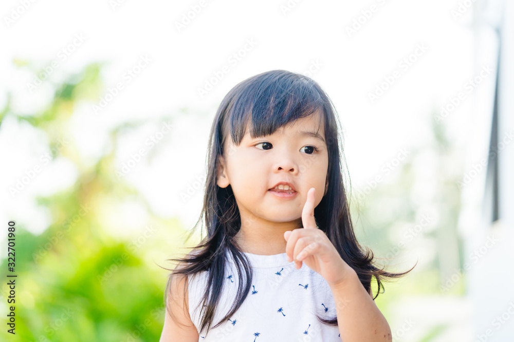 Asian confident child toddler girl in the garden at school shows the index finger upward for thinking and asking question.Very smart schoolgirl thinking and observe around the garden. Asian confident child toddler girl in the garden at school shows the index finger upward for thinking and asking question.Very smart schoolgirl thinking and observe around the garden.