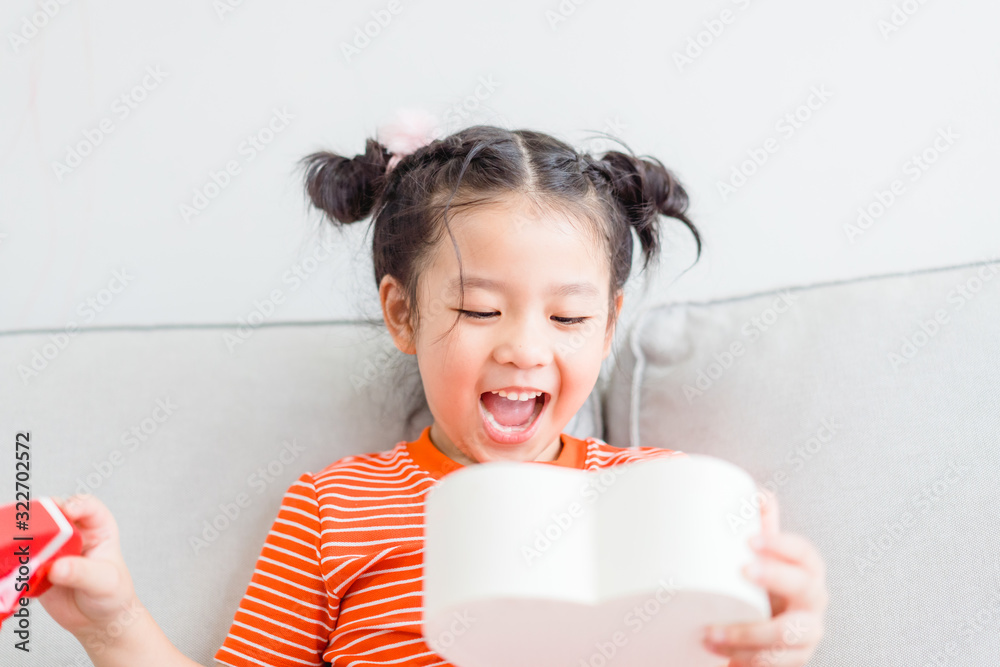 Little asian girl Happy smiley excited face and open red gift box in heart shape in sofa at home on white wall background.Child girl holding gift box in Valentine day, Christmas and New year concept. Little asian girl Happy smiley excited face and open red gift box in heart shape in sofa at home on white wall background.Child girl holding gift box in Valentine day, Christmas and New year concept.