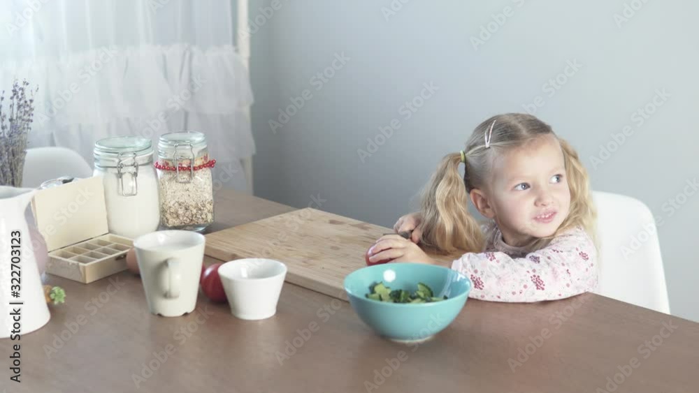 A little girl cuts a tomato for a vegetable salad. Sweet mom's assistant cooks in the kitchen A little girl cuts a tomato for a vegetable salad. Sweet mom's assistant cooks in the kitchen