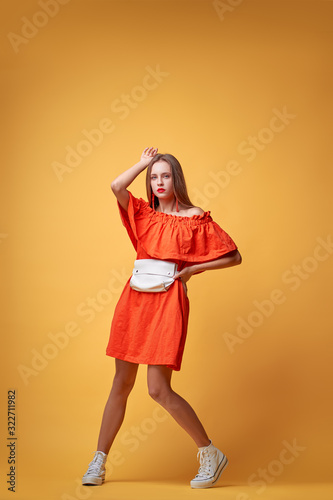 Summer urban fashion. Fun and colorful. Young pretty happy woman in orange dress posing against yellow background.