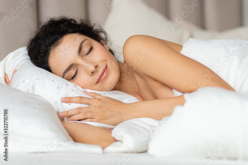Close up face young woman with light smile lying on white pillow. Good sweet dreams and morning sleepiness