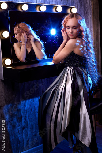 Portrait of young elegant tender blonde teenage girl in black dress in the dressing room near the mirror with flashlights. Caucasian female model with very long hair posing indoors